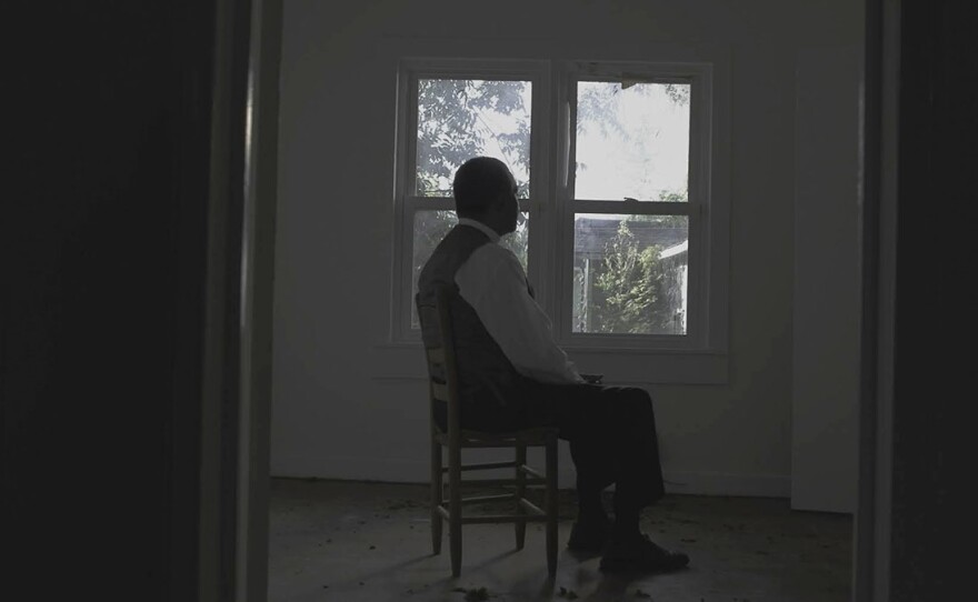 A man is framed through a doorway, sitting in a chair and gazing out of a window with his back facing the viewer. The room is sparsely furnished, with white walls and debris scattered on the floor. The view outside is partially obscured by his presence but glimpses of trees and a house can be seen through the window.