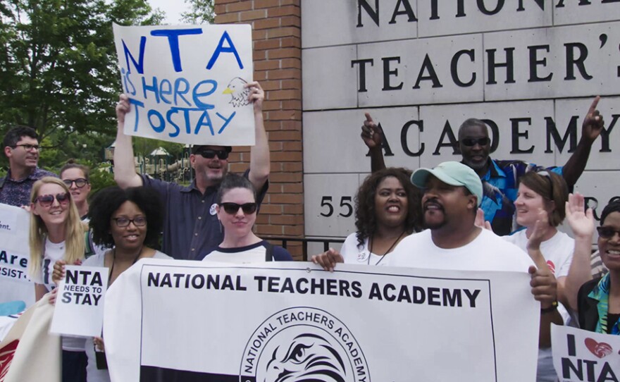 Two protestors hold up a sign saying "National Teachers' Academy" in a crowd of other protestors holding similar signs.