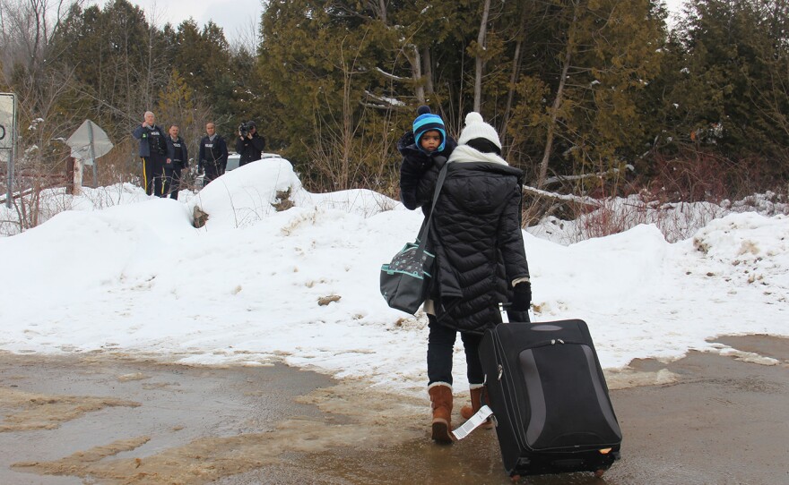 A woman traveling alone with her infant, seeming to understand that she will be arrested, walks toward Canadian police on the far side of the border.