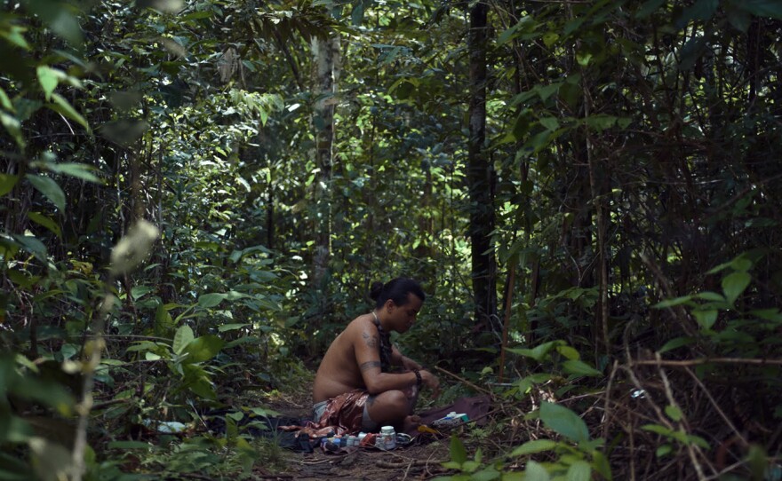 Uýra, a brown-skinned indigenous artist sits crossed legged on the forest ground which is covered with broken sticks and other natural debris. Shirtless and seated in profile in front of a mirror, with arms resting on knees, their skin reveals a smattering of tattoos across the visible right arm. Their head is tilted to the ground as if surveying the various bottles of paint and makeup that are spread across the ground. Uýra's figure is enveloped by the dense forest of green leafy trees that encircle them.