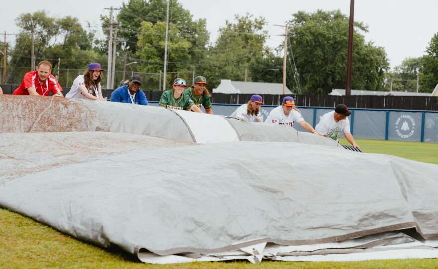 Players with the Boston Hunters and the San Francisco Firebells assist the grounds crew in rolling up the infield tarp prior to the start of the WPBL's inaugural game on Saturday, Aug. 1.