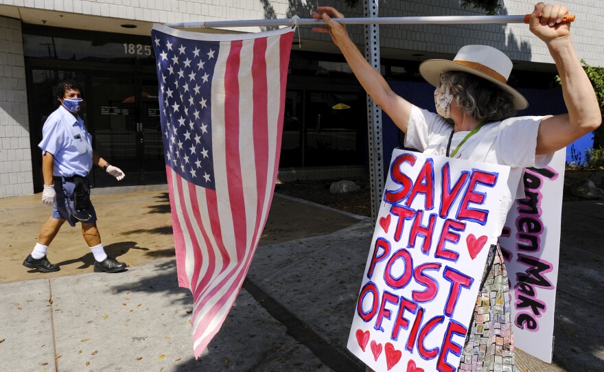 Erica Koesler of Los Angeles demonstrates outside a USPS post office as a postal worker walks by in the background on Saturday. The USPS has warned states coast to coast that it cannot guarantee all ballots cast by mail for the November election will arrive in time to be counted, even if mailed by state deadlines.