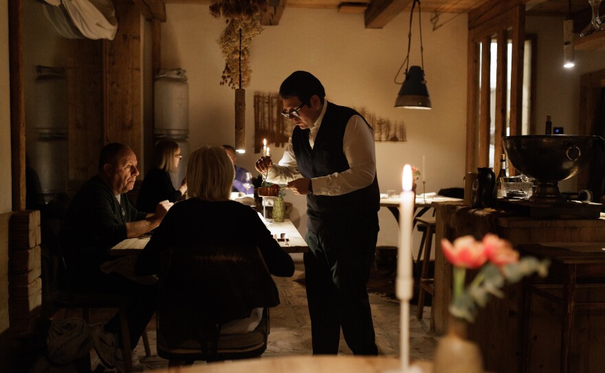 A waiter provides tableside service for guests in the dining room of the Michelin-starred restaurant SanBrite. The establishment, known for its "agricucina" philosophy, combines a refined mountain atmosphere with traditional Cortinese architectural elements, emphasizing a direct connection between local ingredients and high-end hospitality in the heart of the Dolomites.