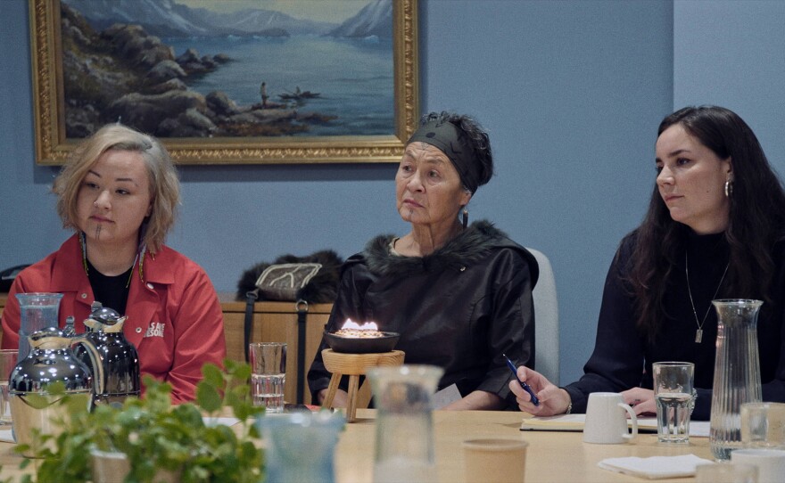 Three Indigenous women sit around a table. The young woman on the left has short blonde hair and a tattooed chin, she wears a red jacket and long earrings. Aaju Peter is in the middle with her short back hair tucked under a black headband and a tattooed chin and forehead. She wears a black fur lined dress and black earrings. On the table something burns in a small iron pot. The woman on the right has long black hair and she wears a black sweater, hoop earrings and a necklace. She holds a pen and notebook open in front of her.