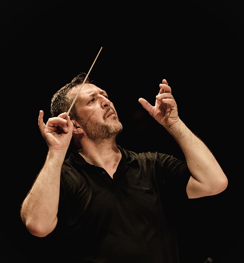 Against a black backdrop, a man conducts an unseen orchestra. He is looking off to the side, with a short beard and mustache, and is wearing a black short-sleeved shirt.