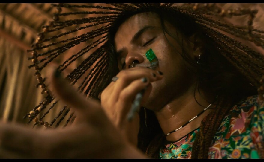 Uýra is pictured under dried palm fronds situated on the right of the frame with head and part of the left shoulder visible. They wear a brightly colored floral pattern top with a thin necklace of black and white beads. With eyes closed and head covered in a hat made of sticks, a paintbrush is held in their right hand capturing the motion of a green line being painted down the left side of their face as the left hand juts out into the foreground just slightly out of focus.