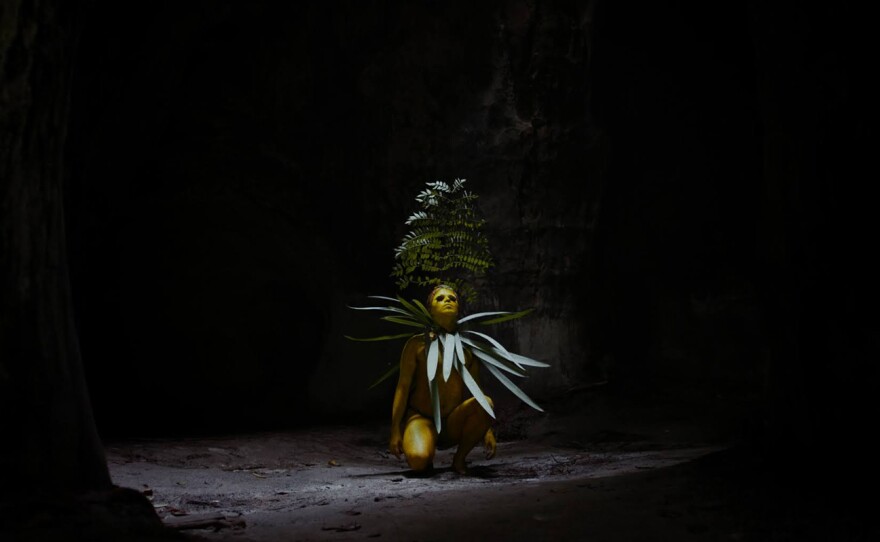 Situated within a dark space a person covered in yellow paint squats down in a pool of light, the right knee is touching the ground, the left knee is bent and arm rest at the side. Their eyes appear closed and painted over with black circles, a large branch covered with leaves juts out from behind the figure to loom overhead, their neck is adorned with long pointed pale green leaves. The ground is covered with grayish dirt, pebbles, sticks and leaves, reflecting the light.