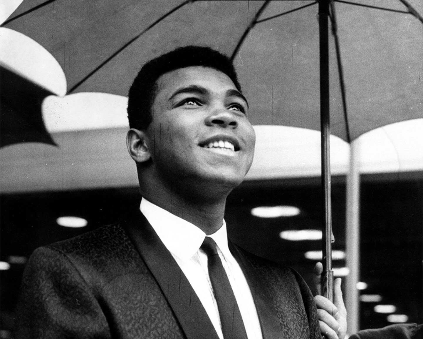 Muhammad Ali smiling as someone holds umbrella for him. He is in Frankfurt to have a title fight to Karl Mildenberger. Frankfurt, Germany. Sept. 2, 1966.
