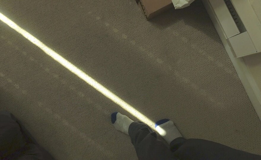 A pair of feet in white and blue socks stand at the end of a bright single line of light cast across the carpet.