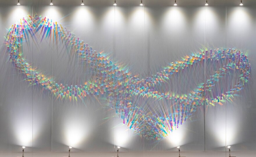 Spotlights shine upwards on a brightly colored wall sculpture consisting of hundreds of tiny pieces of plastic film shaped in a looping, twisting pattern like starlings in flight. The shadows are also colored as the light is refracted and transformed.