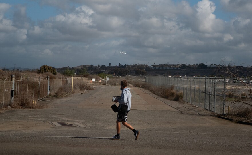A man walks in front of an empty lot looking at a plane in the air in Oceanside, Calif. near Eddie Jones Way and Benet Road, Feb. 11, 2025.