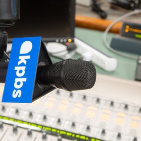 A microphone with a KPBS logo hangs over an audio switchboard in this undated photo.