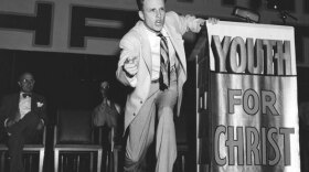 Historical photo of Reverend Billy Graham on stage at a rally for "Youth For Christ." In 1947, at the age of 27, Reverend Billy Graham began traveling across America hoping to spark a spiritual revival with his own conversion story. Believing he could save America by saving souls, he became the country’s most famous preacher of all time.
