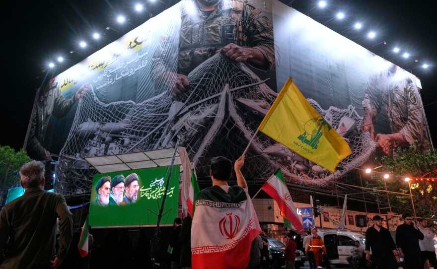An Iranian demonstrator waves a flag of Lebanon's militant Hezbollah group under an anti-U.S. billboard depicting the American aircrafts into the Iranian armed forces fishing net with signs that read in Farsi: "The Strait of Hormuz will remain closed, The entire Persian Gulf is our hunting ground," during a pro-government gathering at Enqelab-e-Eslami, or Islamic Revolution, square in Tehran, Iran, Monday, May 4, 2026.