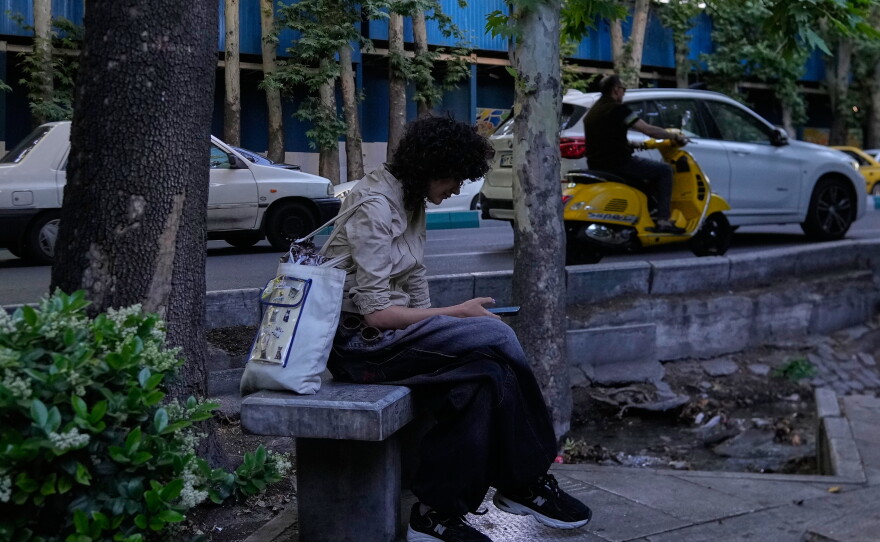 A woman checks her smartphone while sitting on a bench along a sidewalk in northern Tehran, Iran, Tuesday, May 26, 2026.