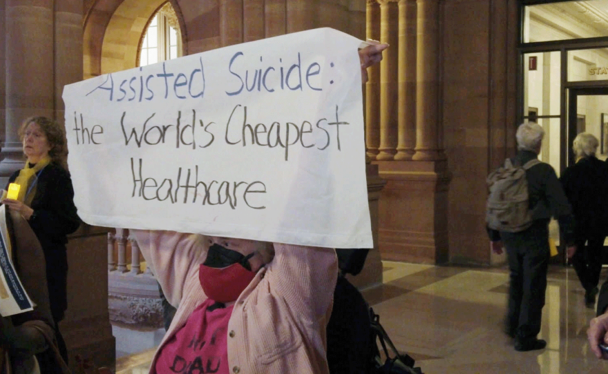 A sign reads "Assisted Suicide: the Worlds Cheapest Heathcare"