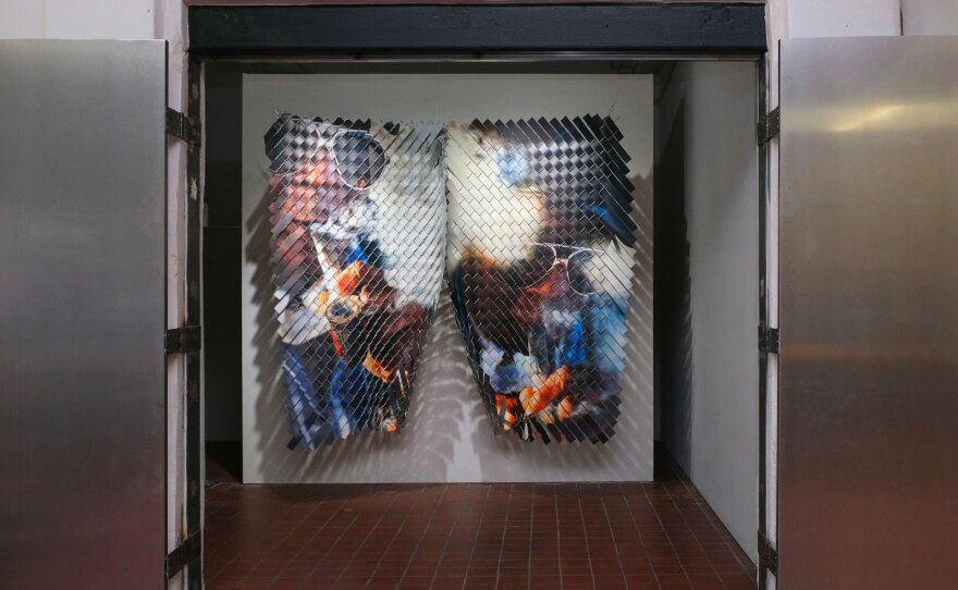 An artwork is shown featuring several fragmented, close up photographs of a man wearing sunglasses and expelling smoke. The pieces are woven through chainlink fence sheets jutting out from the wall.