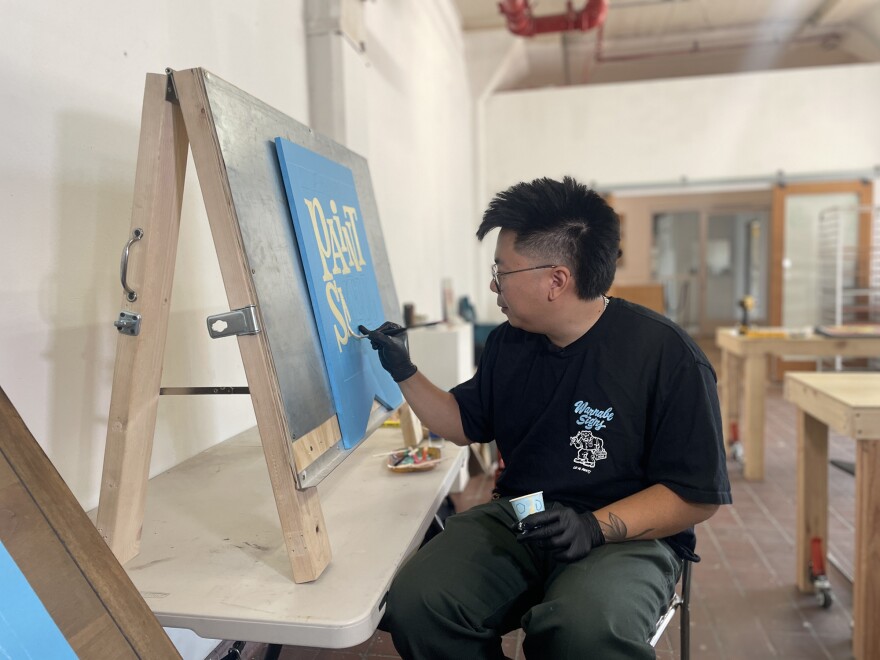 An artist paints a wooden sandwich-board style sign. The sign is painted blue and he is painting cream colored letters that read "PAINT SMELL."