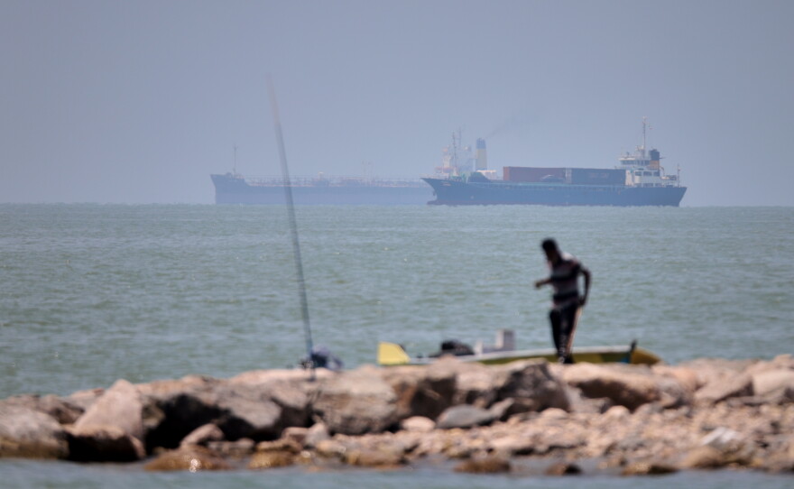 A man stands beside a fishing pole along the shore as cargo ships and commercial vessels are seen in the Strait of Hormuz off Bandar Abbas, Iran, Wednesday, June 17, 2026.