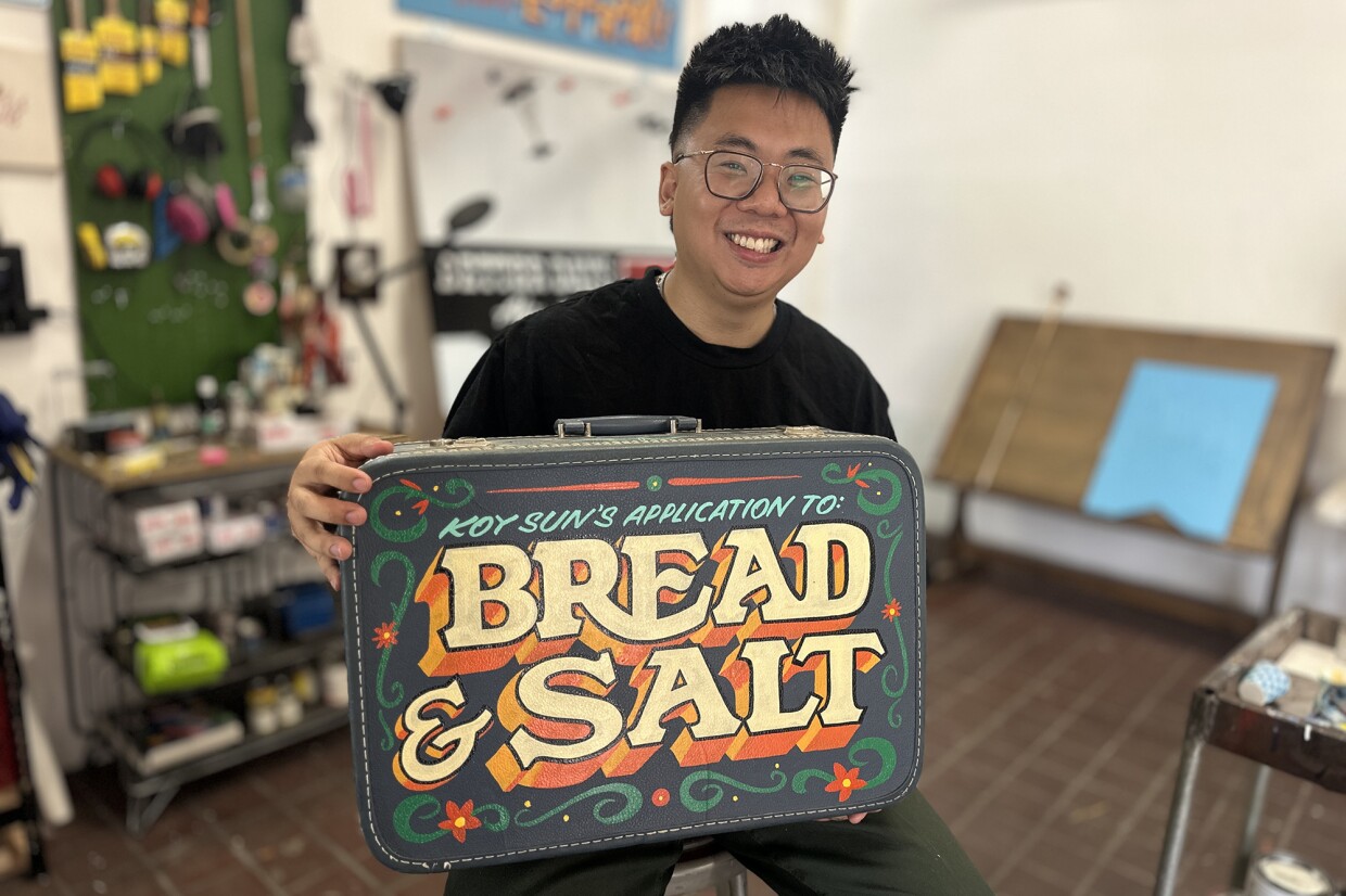Artist Koy Sun is shown in the Impact Artist Residence studio at Bread & Salt in Logan Heights on Oct. 4, 2025. He holds the painted suitcase he used to apply to the program.