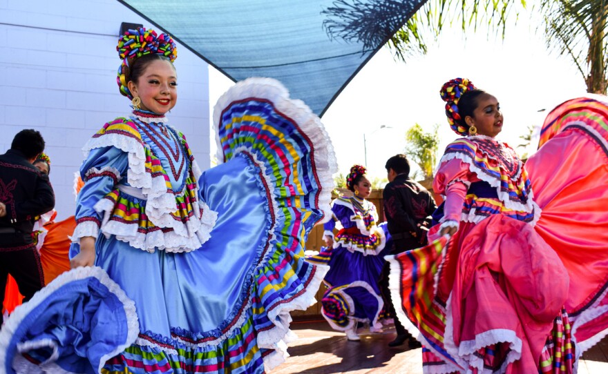 Youth ballet folklorico dancers are shown twirling their dresses. One wears a periwinkle blue-colored dress and the other a bright pink colored dress. The dresses are adorned with multicolor ribbons, and the girls' hair is tied up with multicolor adornments. Other adult dancers dance in the background.