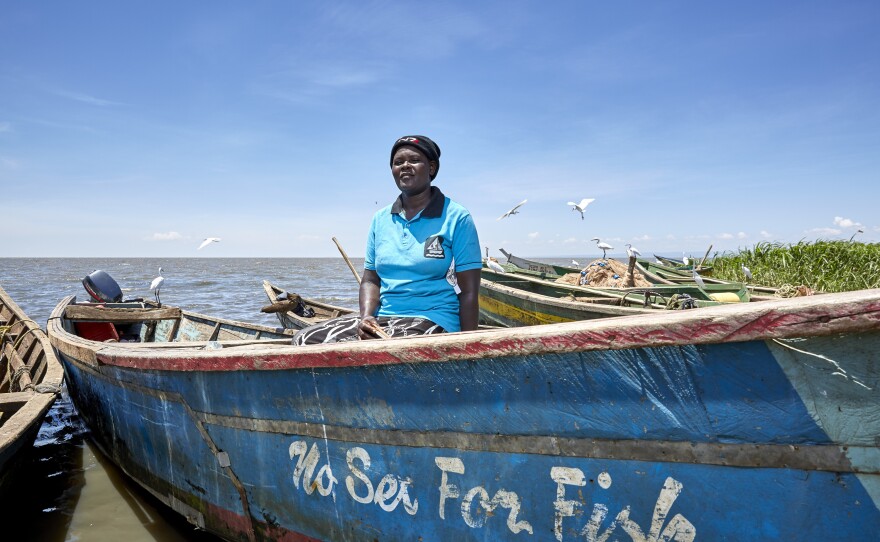 Rebbeccah Atieno stands with one of the boats owned by the No Sex for Fish women. Once a proud boat owner herself, she says she lost her home when Lake Victoria swept through the village; her boat no longer functions. She now earns a living working on rice farms and selling food at a kiosk. A widowed mother of six, she worries about how she'll pay school fees without her fishing income.