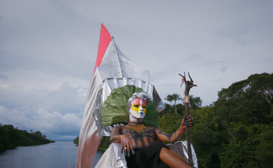 Uýra, a trans-Indigenous artist, reclines on a throne made of various recycled materials as they sail down a river in a performance of "Priscilla: Queen of the Amazon." They wear a vinyl headwrap, with a green rippled fan framing their head, and hold a wooden staff. Their face is painted in vibrant colors of red, yellow, white, and blue, and their body is adorned with geometric tattoos.