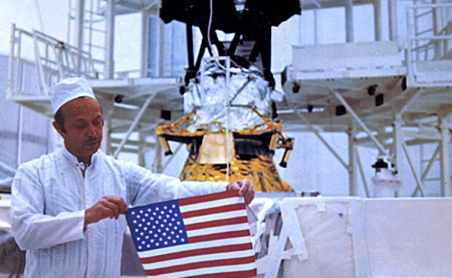 A technician holds a U.S. flag over a gold record containing data from Earth, with a Voyager spacecraft in the background.
