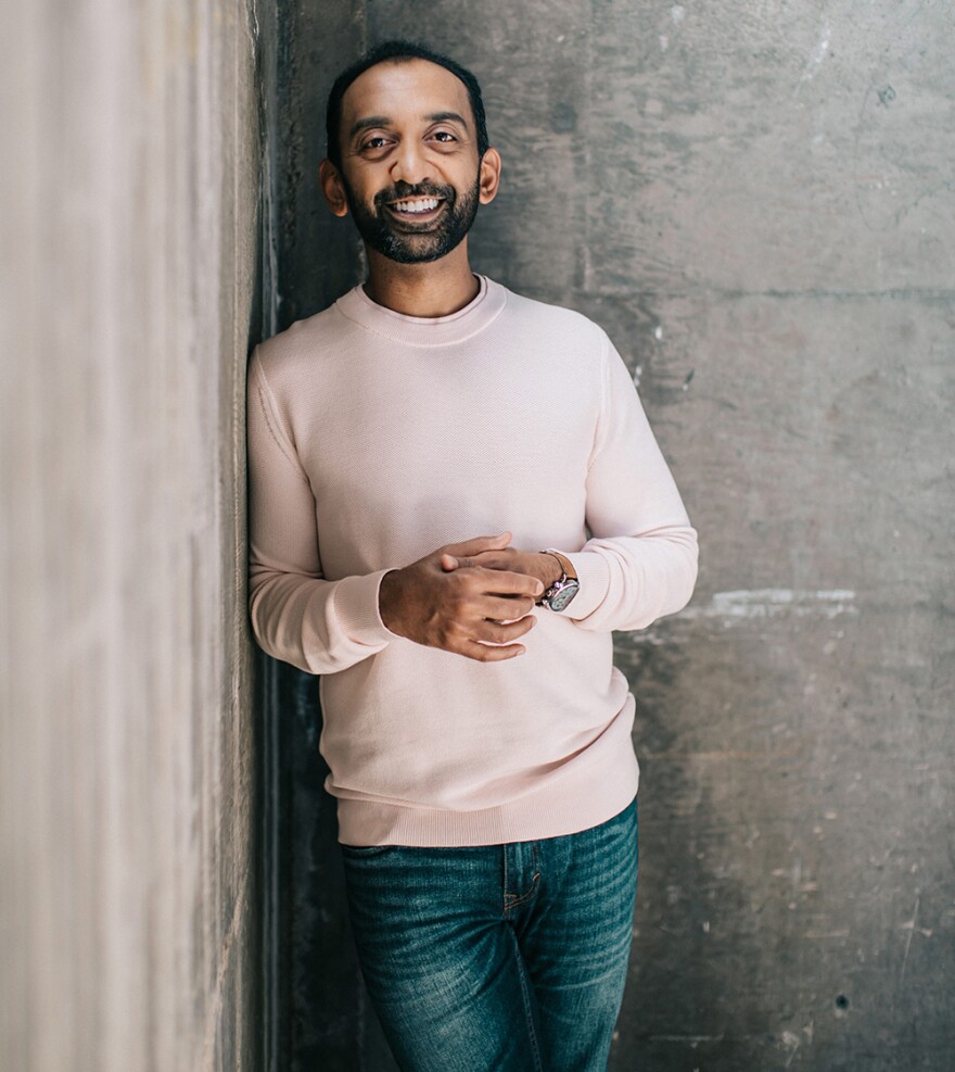 Sameer Patel, conductor of the La Jolla Symphony and Chorus and artistic director of San Diego Youth Symphony, is shown in an undated photo. He is wearing an off-white sweater and jeans and is leaning against a concrete wall. He is smiling and looking at the camera.