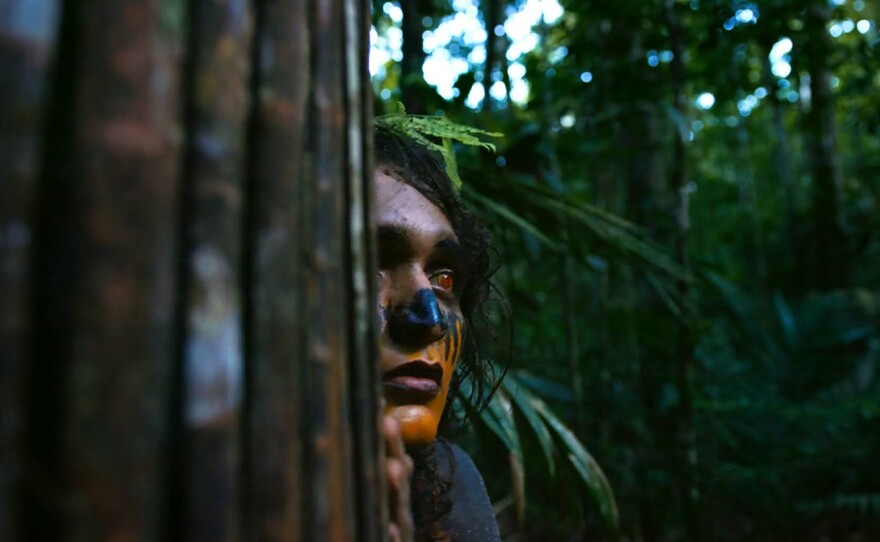 A face peers out from behind a dark brown tree trunk with finger tips touching the tree. Though the face is partially obscurred we see a left eye which is beset with a red contact lens gazeing out towards the right side of the frame, their nose is painted black and closed lips colored a dark red, cheek and chin shaded orange. A dense mass of dark green foiliage with some light streaming in surrounds the face.