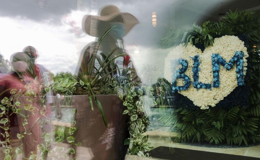 People are reflected in a window near a flower arrangement that includes the acronym for Black Lives Matter as they wait in line to attend the public viewing for George Floyd at the Fountain of Praise church on June 8, 2020 in Houston.