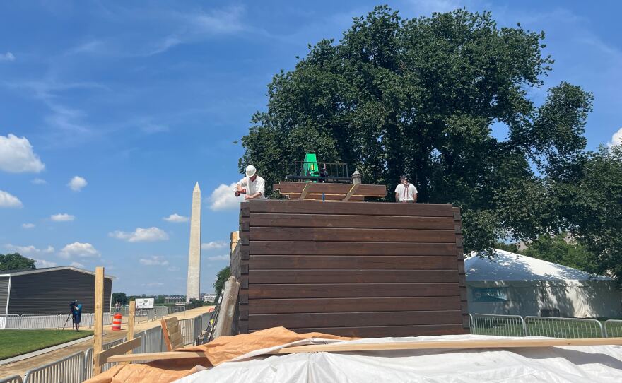 Workers assemble a home resembling an instant log cabin on the National Mall in Washington, D.C., with the Washington Monument behind them.