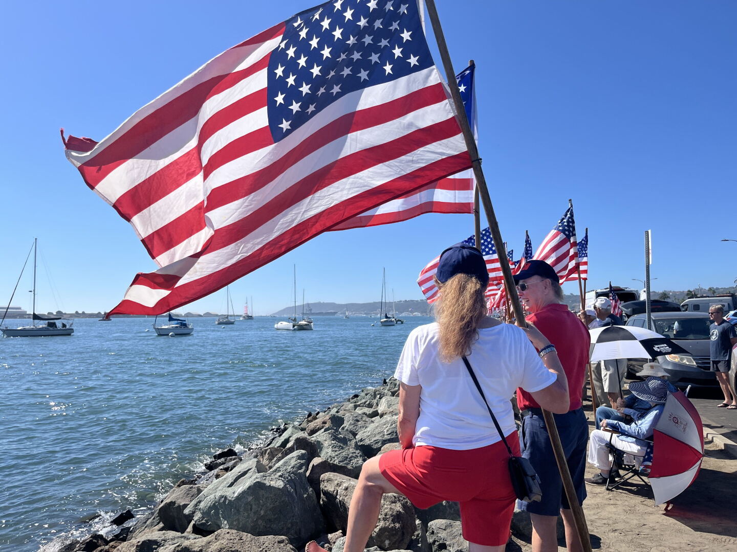People gather with American flags along Shelter Island's shoreline ahead of the USS Lincoln's arrival on Oct. 8, 2026.