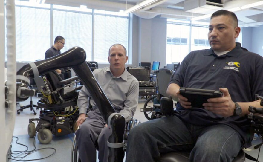 Rory Cooper, PhD, (left) looks on as a researcher at the Human Engineering Research Laboratories in Pittsburgh, Pa. tests a wheelchair fitted with a robotic arm. Cooper, a disabled veteran, co-founded the labs with the mission to improve mobility for all people with disabilities.
