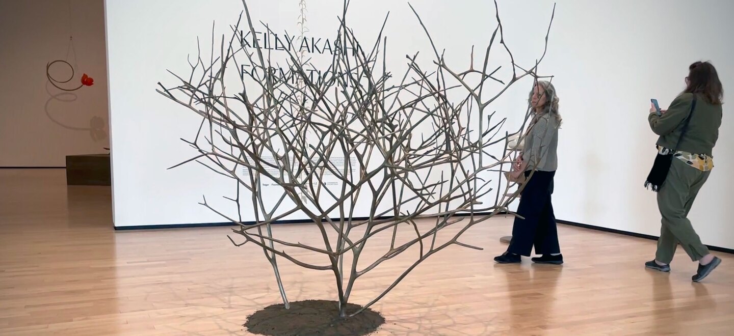 A tree-like sculpture is shown on a wooden museum floor, atop a circle of dirt. Two women walk past the sculpture.