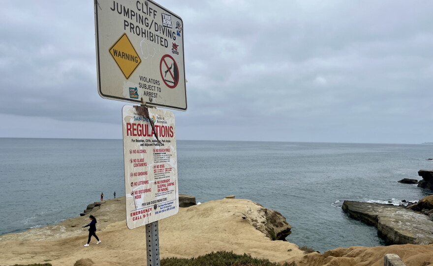 A sign signaling the prohibition of cliff diving stands in the parking lot as people walk near Pappys Point in Point Loma, Aug. 5, 2026.