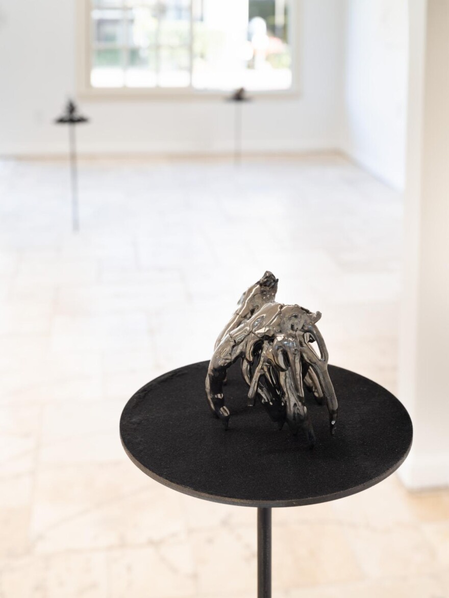 A shiny metallic sculpture sits on a black pedestal. The sculpture looks like a sea creature made of molten metal. In the background, blurry, are two other similar black pedestals with unfocused, indistinguishable small sculptures