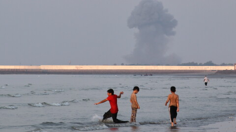 Three boys play in the shallow waters of the Strait of Hormuz, as a plume of smoke rises from an explosion in the background, off Bandar Abbas, Iran, Monday.