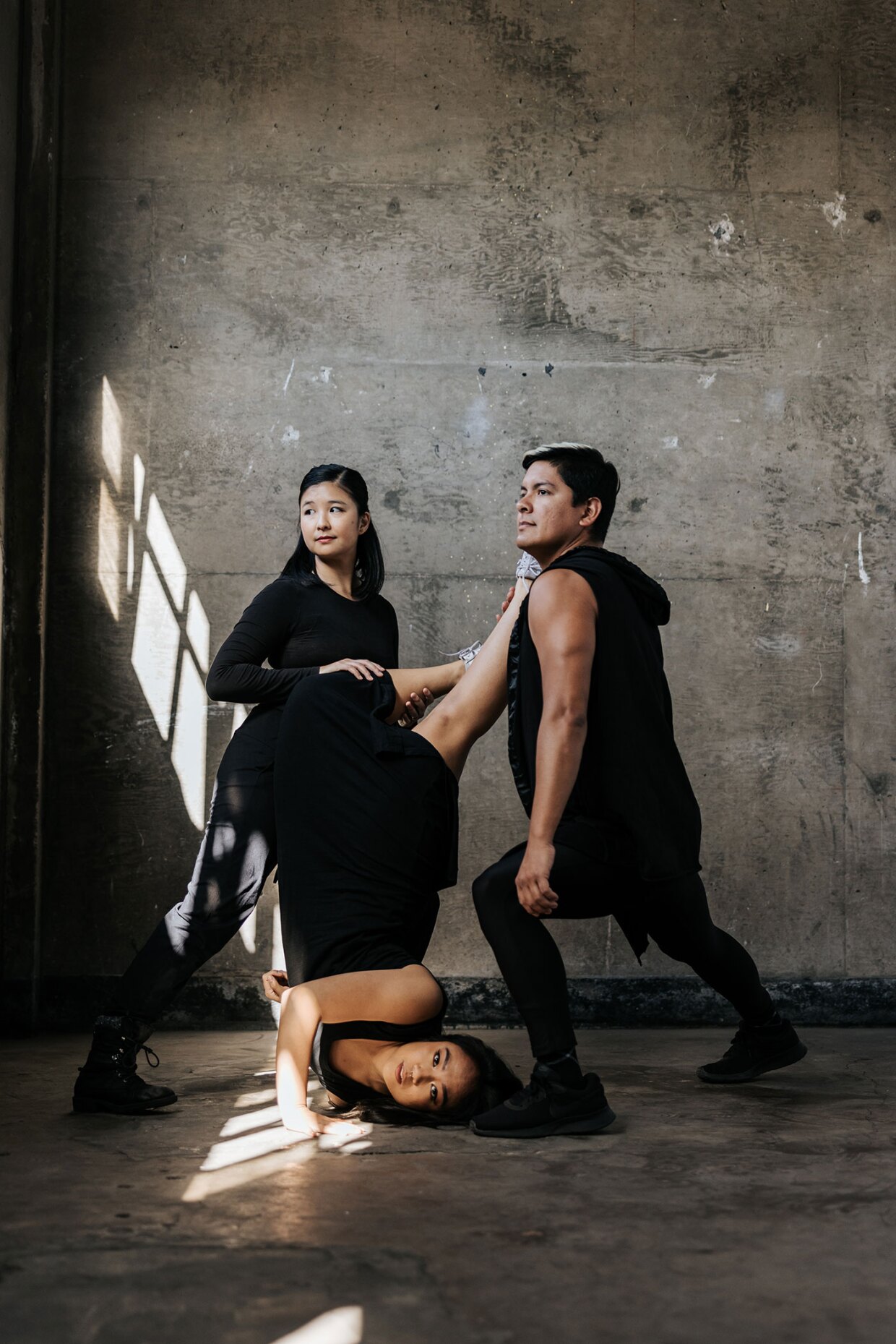 Three dancers dressed in black perform in a concrete studio.