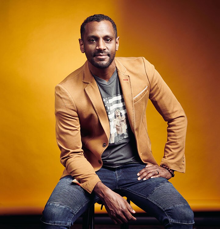 Composer Carlos Simon is shown against a bright orange backdrop wearing blue jeans, a beige blazer and a t-shirt. He is lightly smiling and looking at the camera. He has medium-to-dark brown skin and black hair and a close-shaved beard.