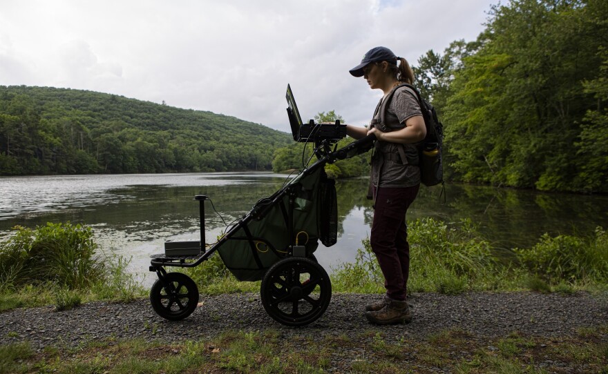 Standing on a gravel path with a lake behind her, Lillian Mullock checks the laptop atop the HETAP device for recent data points as part of a training session at the Delaware Water Gap.
