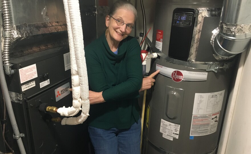 Jane Stackhouse points to the electric heat pump (left) that replaced a gas furnace and to a more efficient electric water heater (right). Her utility bills are about the same, but now she also has air conditioning for extra warm days made worse by climate change.