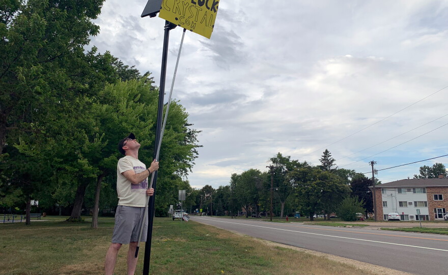Brady O'Rourke in Crystal, Minn., holds a sign he made to block the view of a Flock camera, which tracks vehicles driving along the road. It's one of several actions Americans are taking to oppose this police surveillance technology.