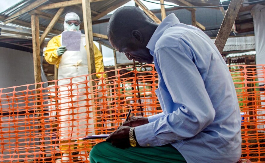 A health worker shouts out details about a case from inside the fence around the isolation area at the Kailahun treatment center. A colleague outside the fence writes them on the patient's chart.
