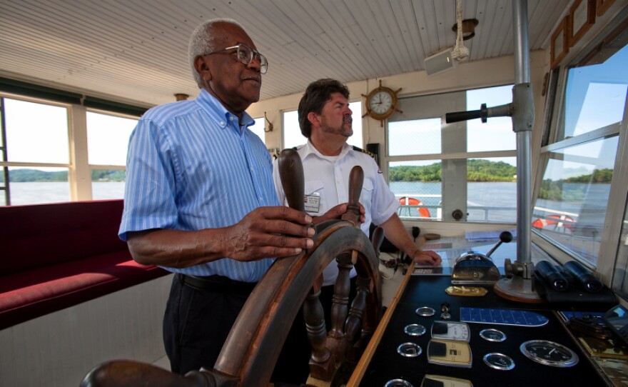 Sir Trevor McDonald takes the wheel of a riverboat with the help of the captain on the Mississippi River. 