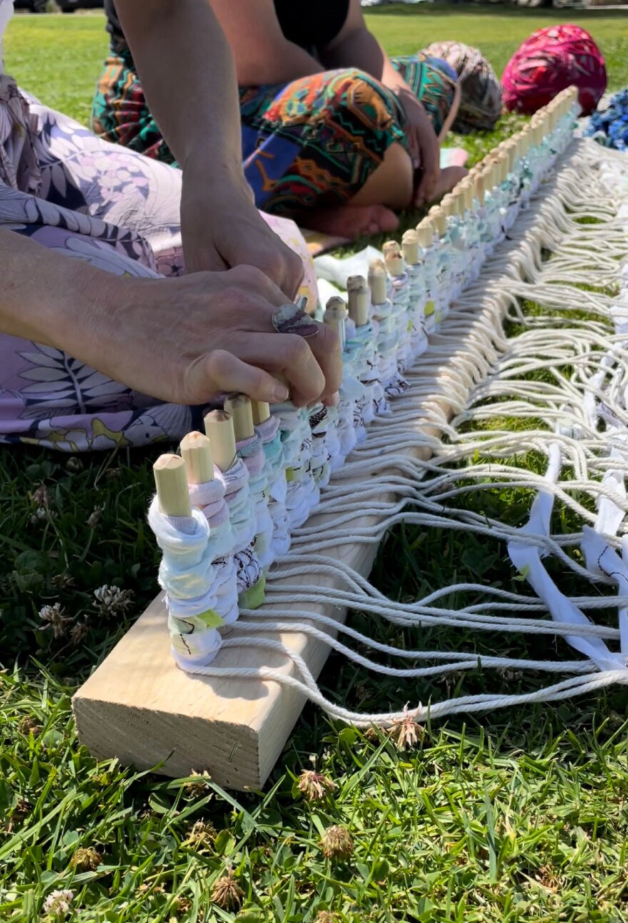 a homemade loom holds strips of clothing, while two artists loop the fabric onto the loom