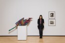 MCASD senior curator Jill Dawsey is shown standing near art from "For Dear Life: Art, Medicine, and Disability" in an undated photo. The works shown are Judith Scott's 1997 mixed media and string sculpture, "Untitled," and Jay DeFeo's 1989 series of graphite on paper, "Untitled (Reflections of Africa series)." Jill is wearing black pants and boots, a pink shirt, and a black cardigan. She has her hands tucked in her pockets, and she is looking at the camera and smiling slightly. She has brown hair and light skin.