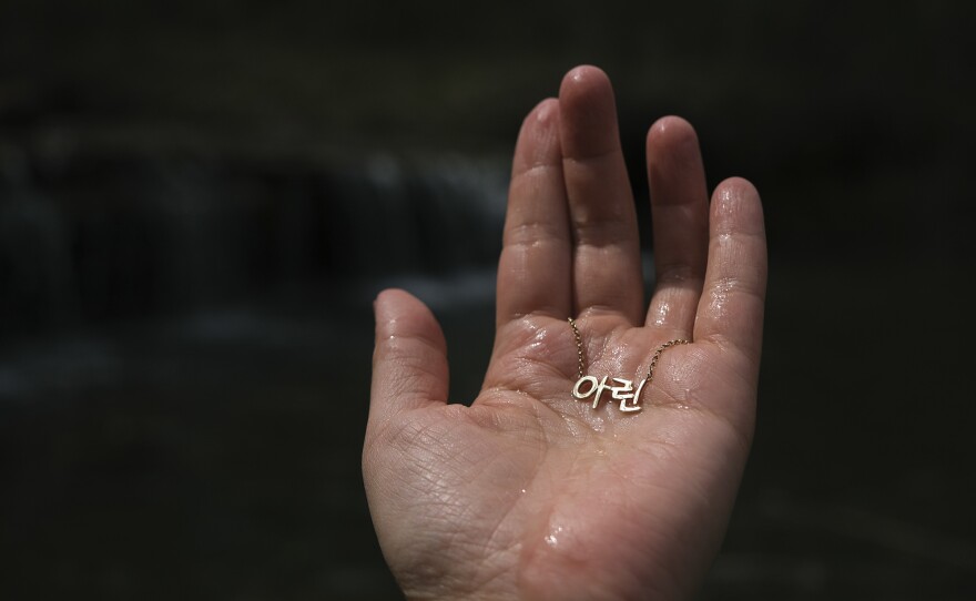 In her conception dream, Young Ok Na, the photographer's mother, holds up jewels from the stream to the light. She thinks of the Korean character 아<em> </em> (pronounced 'Ah'), which stands for beauty, and <em>린</em><em> </em> (pronounced 'Lhin'), which is the sparkle of a jade. In an interpretation of that moment, Arin Yoon holds up her Korean name necklace to the sunlight.
