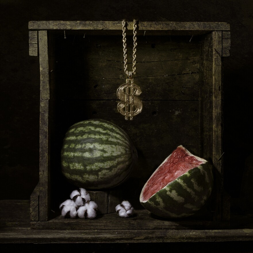 An extremely oversized golden dollar sign necklace is suspended from a wooden frame, above a tableaux consisting of a whole watermelon, a sliced open wedge of watermelon, and some small white flowers. 