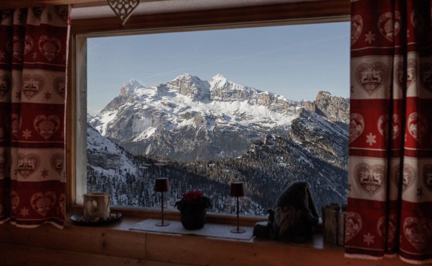 The snow-capped peaks of the Tofane massif are framed through a window of a rifugio, a kind of traditional mountain hut, decorated with typical heart-patterned Ampezzo textiles. These high-altitude lodgings serve as essential rest areas for skiers and hikers, offering a blend of rustic hospitality and panoramic views that define the winter experience in the Dolomites.
